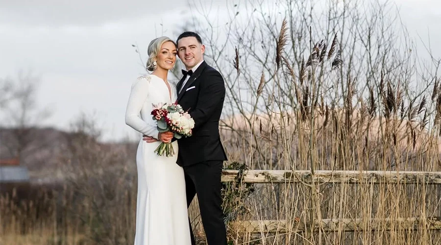 A newlywed couple standing outdoors, with the bride holding a bouquet, in front of tall grasses and trees during daytime.