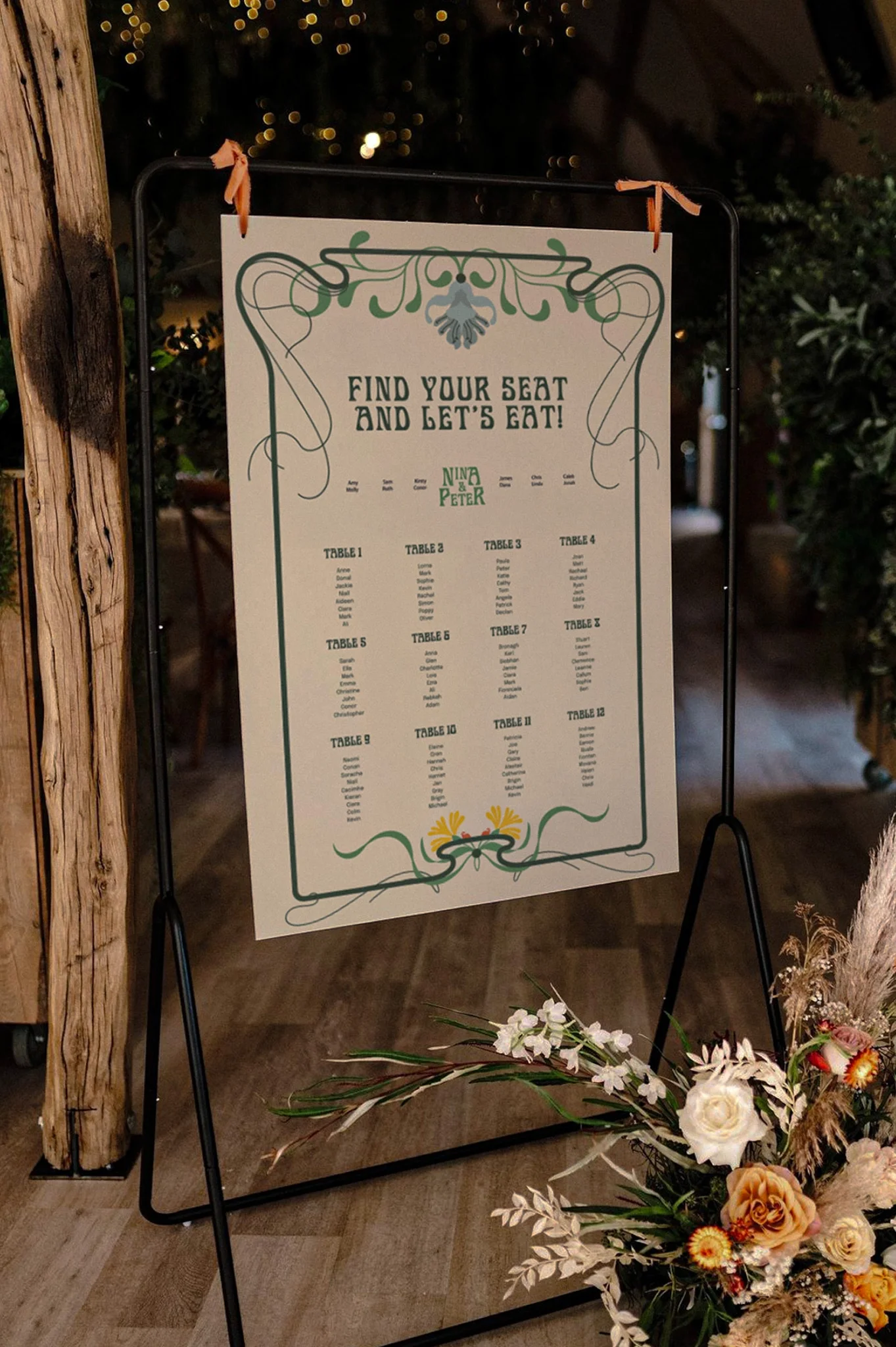 A wedding seating chart with the heading 'Find your seat and let's eat!'. It lists tables and guest names, decorated with floral and swirly designs, placed on a black metal stand with a floral arrangement at the base, indoors with warm ambient lighti