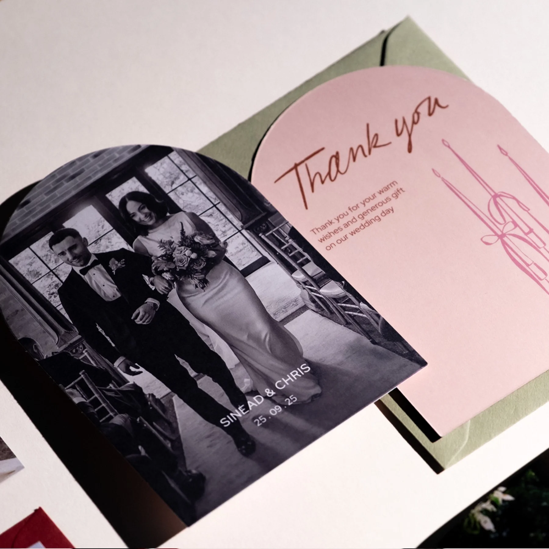 A wedding thank you card featuring a black and white photo of the bride and groom walking arm in arm, with the bride holding a bouquet. The card is placed on a green envelope, and another pink card with "Thank you" written in cursive is partially vis