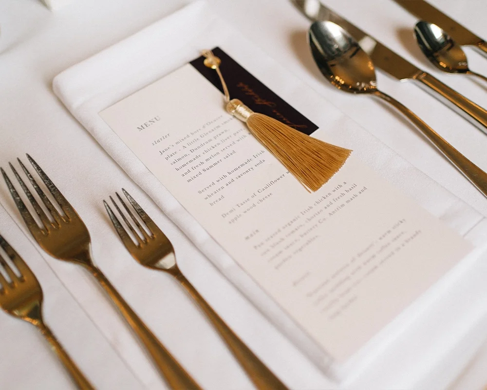 Elegant table setting with three gold forks on the left, three gold spoons on the right, and a handwritten waiter's note with a tassel on top, placed on a white napkin and a printed menu on a white tablecloth.