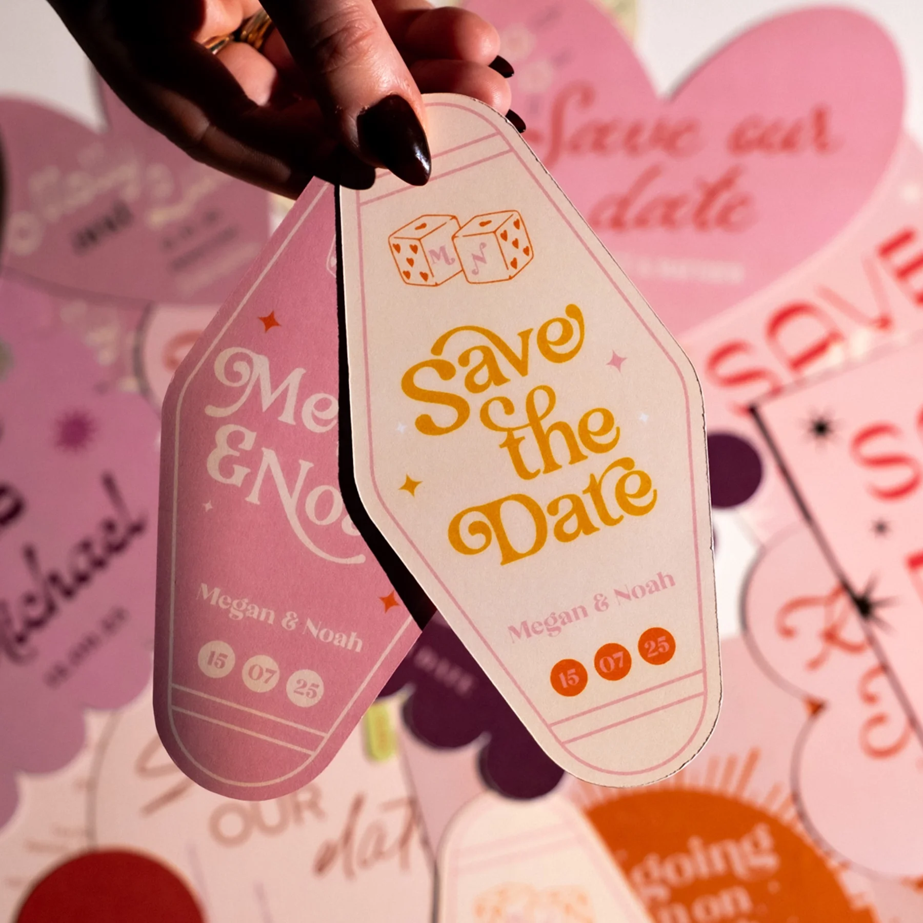 A pink and white Save the Date tag with the names Megan and Noah, a date (15 07 25), and an illustration of two dice with hearts.