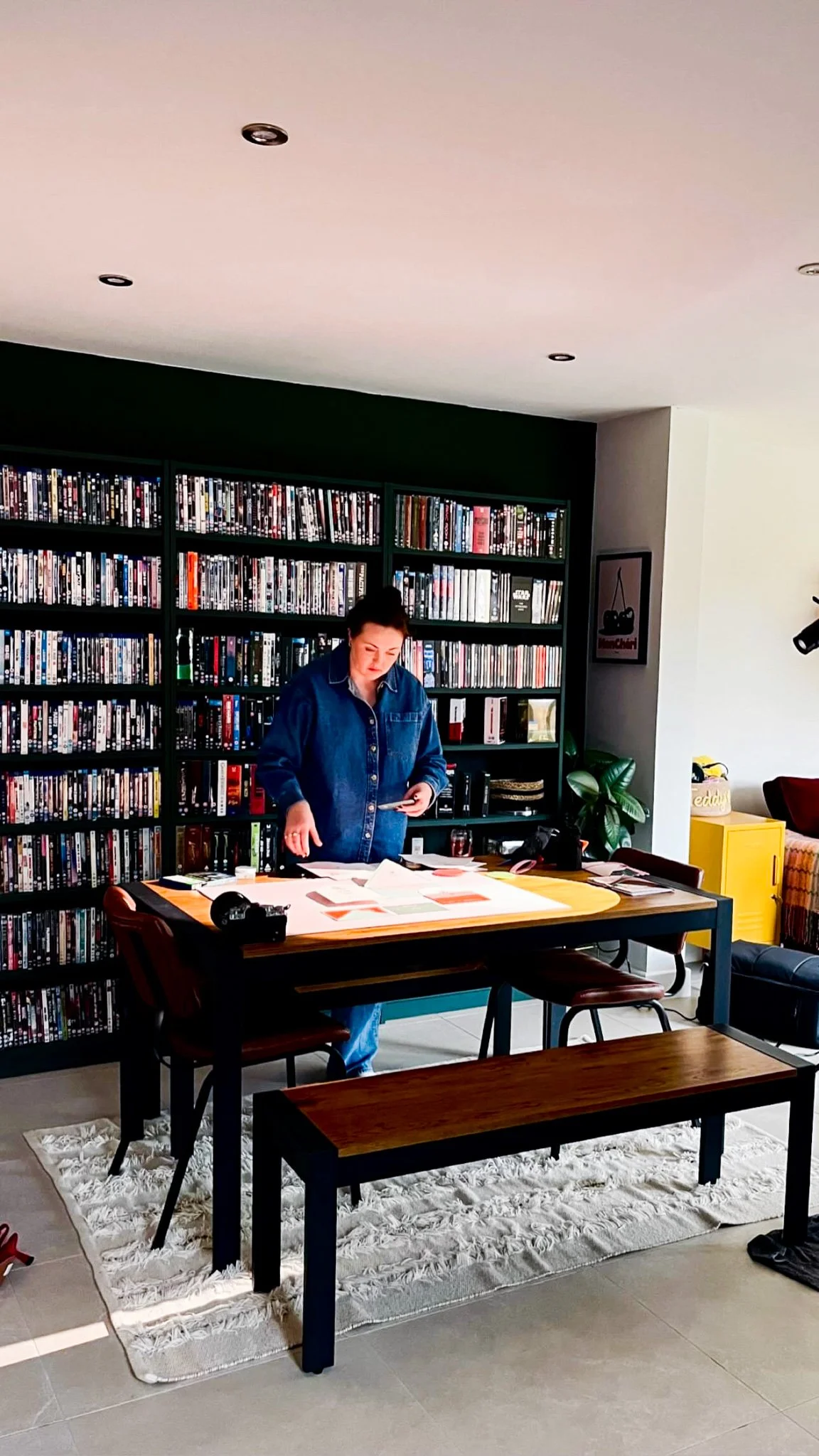Woman in denim jacket standing at a table with papers, in front of a large bookshelf filled with DVDs in a living room.