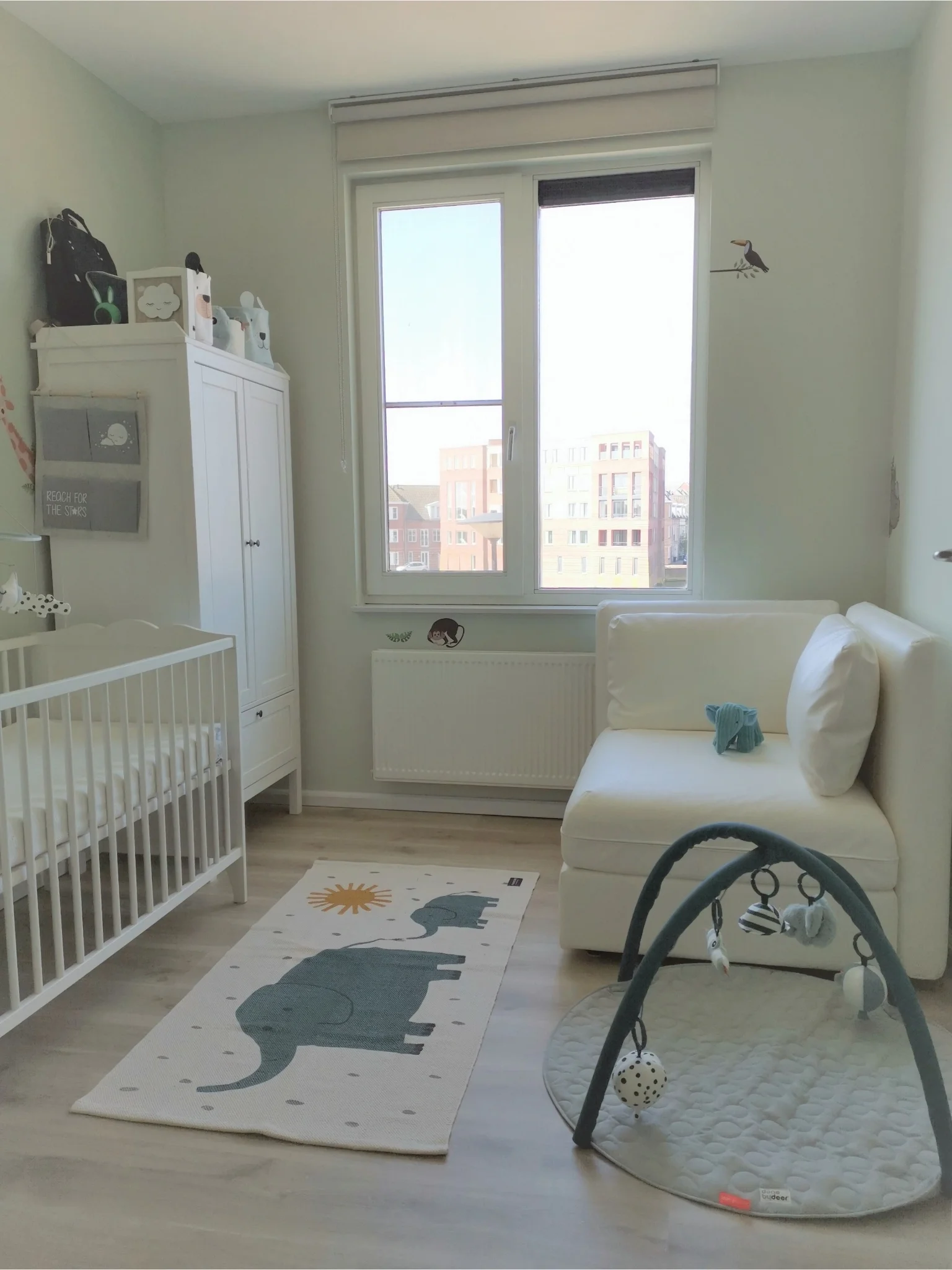 Lichte babykamer met wit ledikant en kledingkast, een bankje bij het raam en een speelkleed met olifant op de vloer.