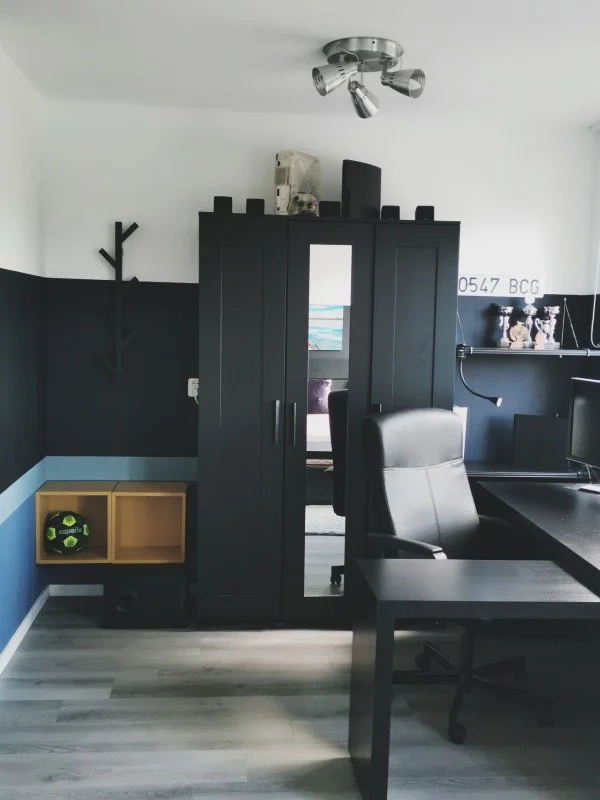 Stoere jongensslaapkamer met zwarte kledingkast, modern bureau, ergonomische bureaustoel en oker hangkastjes als speels kleuraccent in een donkerblauw interieur