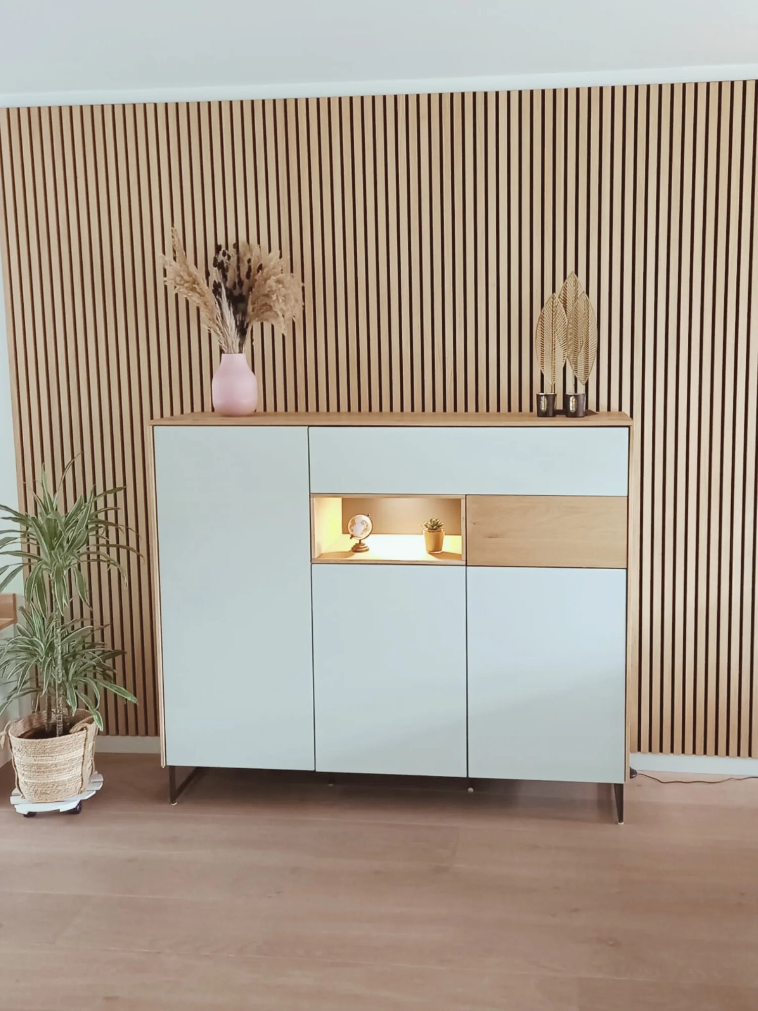Houten lattenwand met modern dressoir in zachte groentint en een warm verlichte nis voor accessoires. Rustige basis, natuurlijke materialen en een strak lijnenspel zorgen voor een stijlvolle, gezellige en casa met karakter.  Interieuradvies, interieu