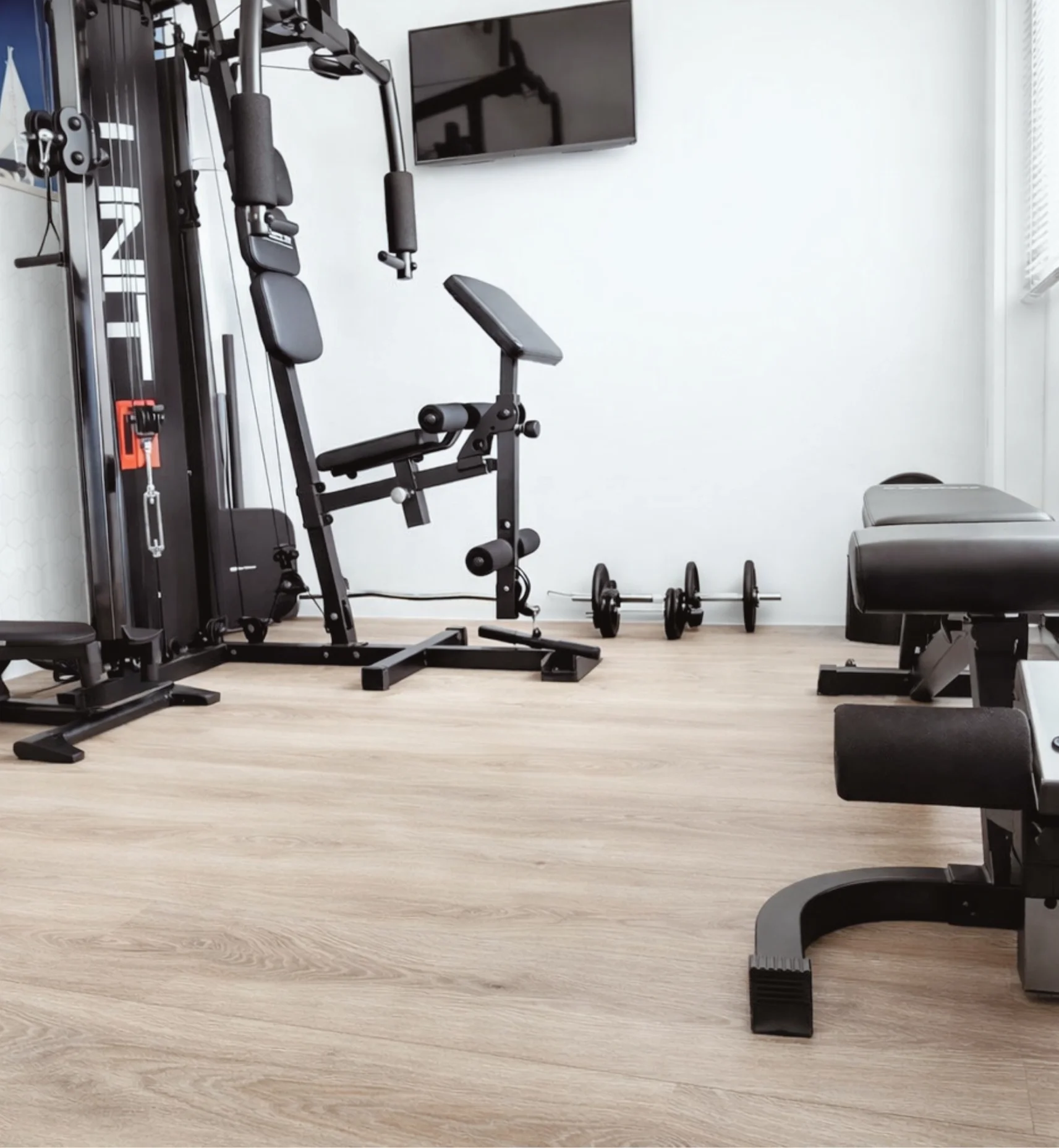 Moderne home gym met krachtstation, trainingsbank en halterset, waar lichte houttinten en een rustige basis zorgen voor een comfortabele en functionele sportruimte aan huis – En-Casa interieuradvies en styling in Heemskerk.