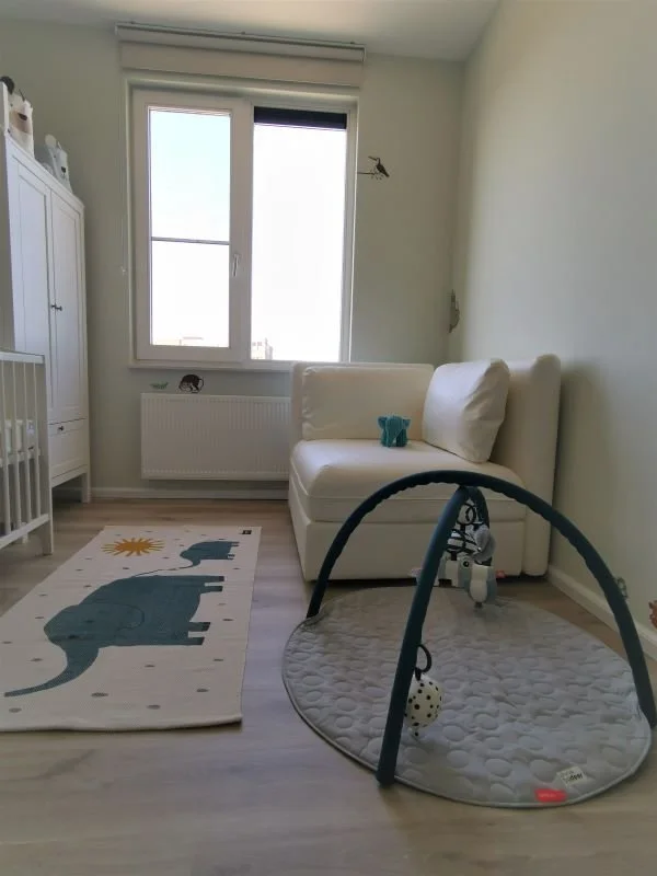 Rustige, lichte babykamer met zachte groentint op de muren en een houtlook vloer. Links staan een wit ledikant en een hoge witte kast. Het grote raam met rolgordijn brengt veel daglicht binnen, met een radiator eronder. Rechts staat een compact wit b