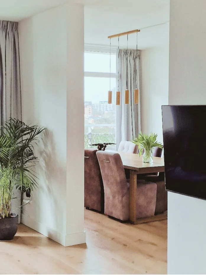 Doorkijk naar de eethoek vanuit de woonkamer: open indeling met veel licht, warme houtvloer, planten en zachte tinten voor een rustige sfeer.  Interieuradvies, interieurontwerp en styling door En-Casa Interiors in Noord-Holland.