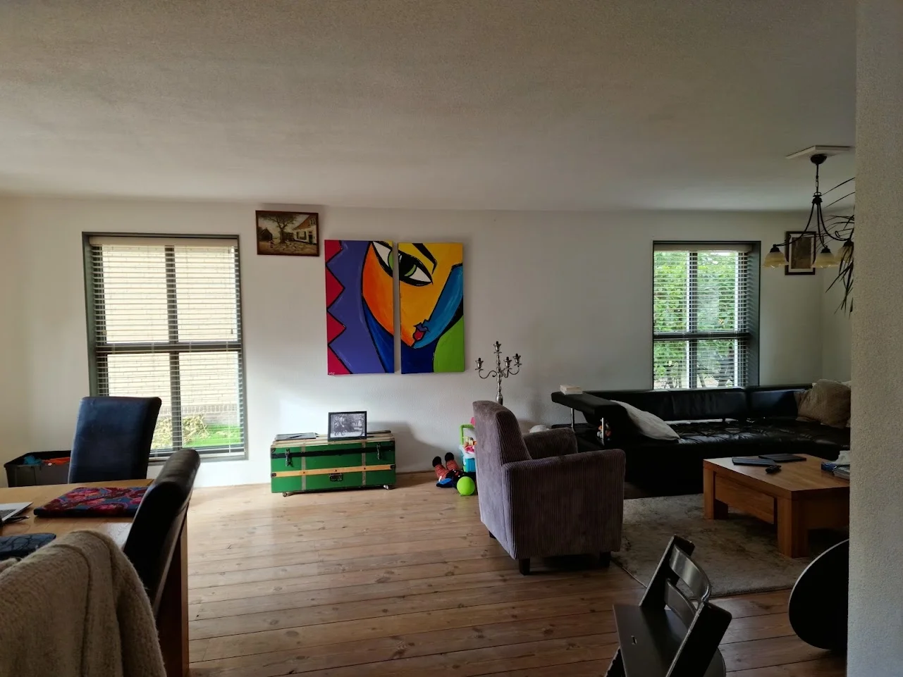 Woonkamer in before-situatie met houten vloer, zwarte leren bank, paarse fauteuil, houten salontafel en kleurrijk schilderij aan de muur. Grote ramen met jaloezieën, ruimte eethoek  met een mix van zit- en eethoek.