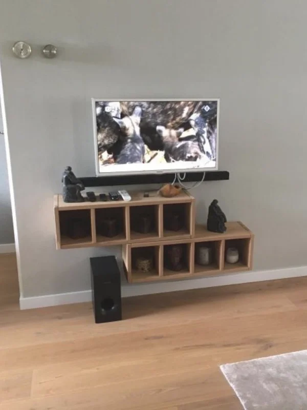 Deze foto toont een rustige tv-hoek uit de woonkamer & eetkamer make-over, met een strak opgehangen televisie en een warm houten tv-meubel met open vakken. De natuurlijke houttint geeft direct warmte aan de lichte wand, terwijl de minimalistische sty