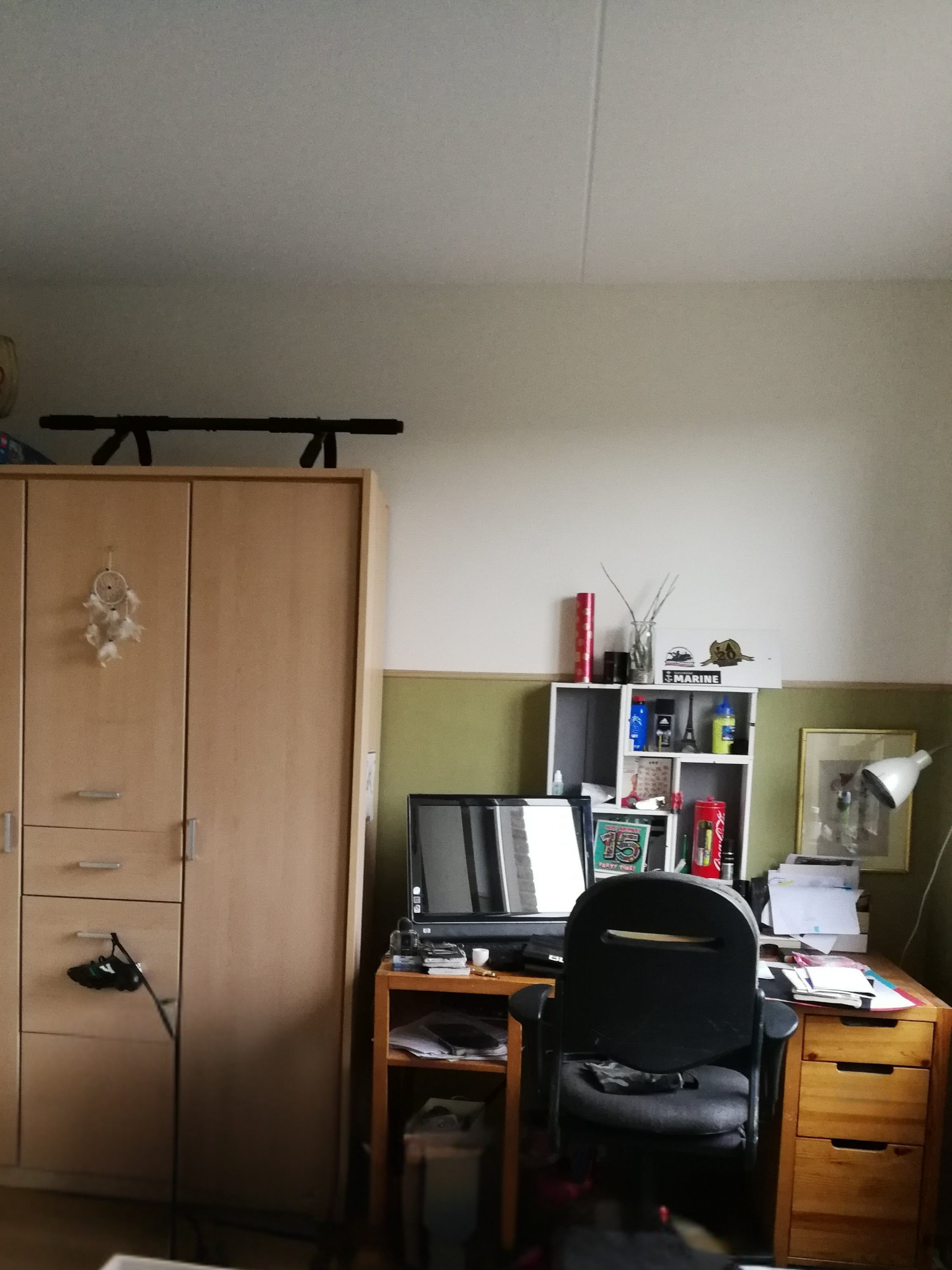 Voor-situatie van de werk-/slaapkamer: een functionele plek met een bureau, veel losse spullen en verschillende opbergelementen bij elkaar. De hoge kast en het bureau zorgen voor de basis, maar door de open vakken en items op het werkblad oogt de hoe