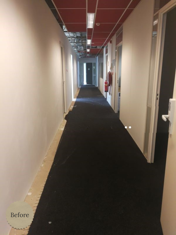 Voor de verbouwing: lange kantoor-/bedrijfsgang met witte wanden, donkere tijdelijke vloerbedekking en zichtbare installaties aan het plafond. Ideale “before” foto voor een renovatieproject van een moderne, warmere werkomgeving.