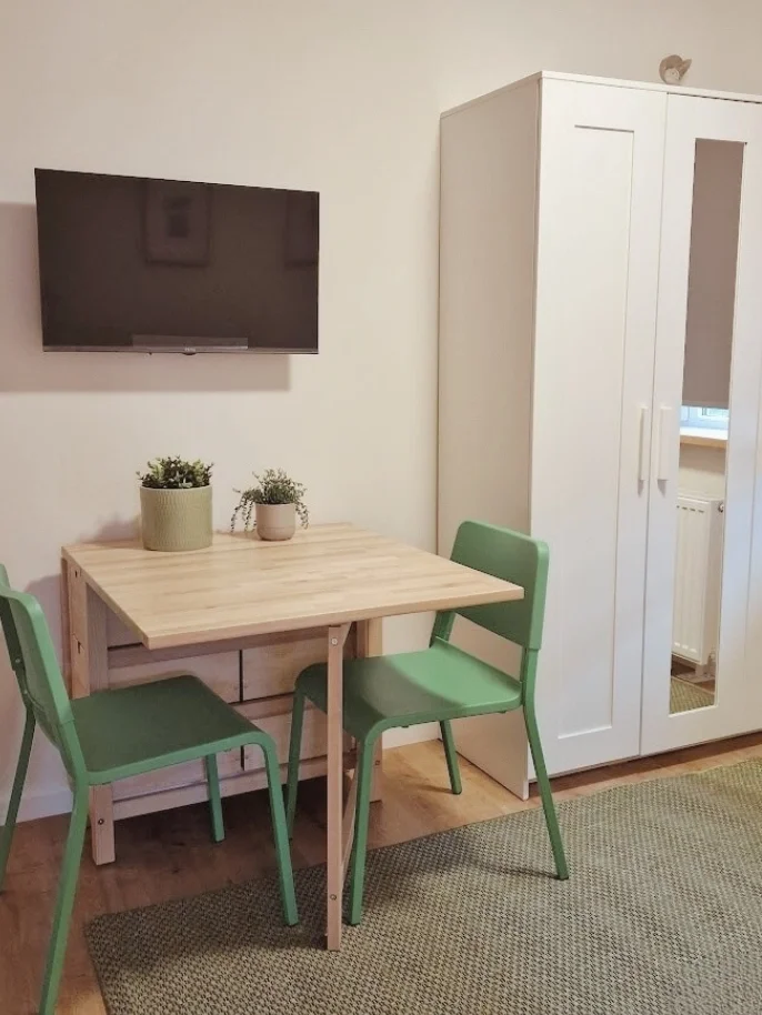Een compacte eethoek met houten tafel en groene stoelen, aangevuld met een wand-tv en een lichte opbergkast — een praktische, gezellige plek om te eten, werken of even bij te komen, met een frisse touch groen voor extra en-casa sfeer.