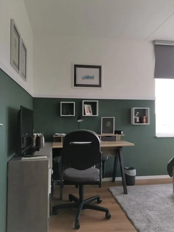 Deze werkplek is stoer en functioneel tegelijk. En-Casa combineerde warm hout met zwart accent en groen behang in de lambrisering, zodat alles één geheel vormt met de rest van de kamer. De wandkubussen zorgen voor persoonlijke styling zonder dat het 