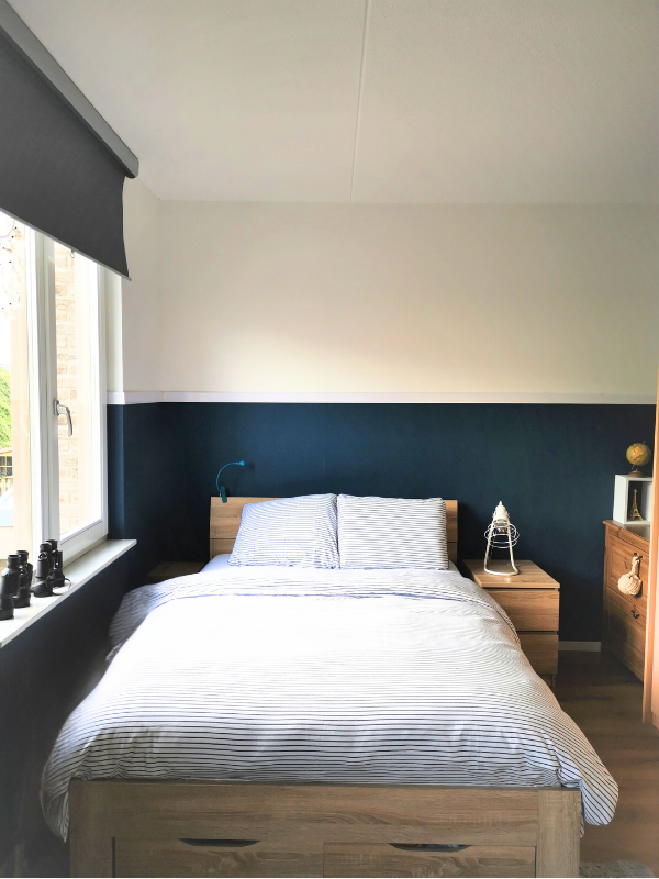 Een rustige, lichte slaapkamer met een houten bed als warme basis en een diepblauwe accentwand die meteen sfeer en diepte geeft. Het frisse, lichte beddengoed houdt het geheel luchtig, terwijl het contrast met de donkere wand zorgt voor een gebalance