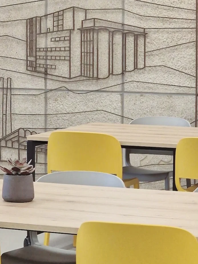 Sfeervolle, moderne werk- of lunchruimte met lange houten tafels, gele stoelen en een industriële stalen wand, afgewerkt met een grote schetswand als eyecatcher—ideale inspiratie voor een warme, creatieve en functionele inrichting. Interieuradvies, o