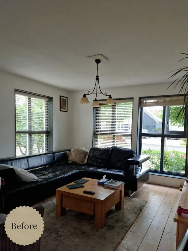 Woonkamer in before-situatie met zwarte leren hoekbank, houten salontafel op een licht vloerkleed, grote ramen met jaloezieën en veel daglicht. De ruimte heeft een rustige, neutrale basis met houten vloer en uitzicht op groen buiten.