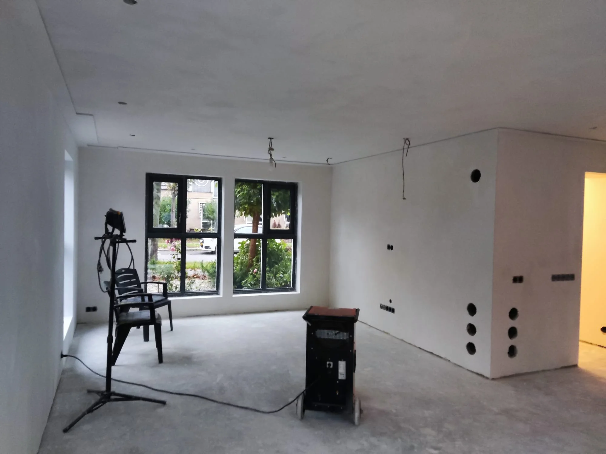 “Woonruimte tijdens verbouwing met gestucte witte wanden, onafgewerkte betonnen vloer en zichtbare elektra in plafond en wanden."