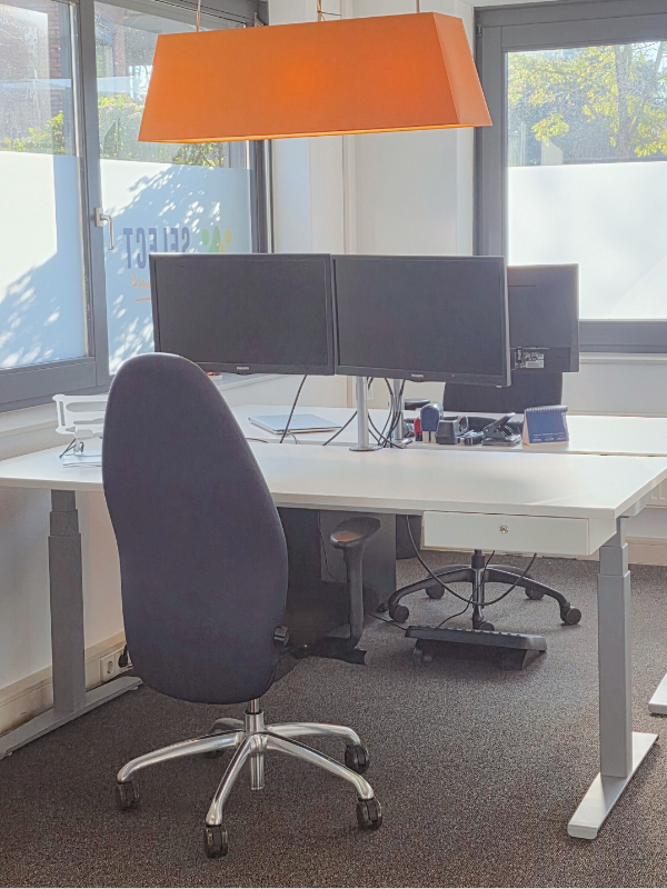 “Werkplek met wit bureau, dubbele monitor en oranje hanglamp, met veel daglicht.”