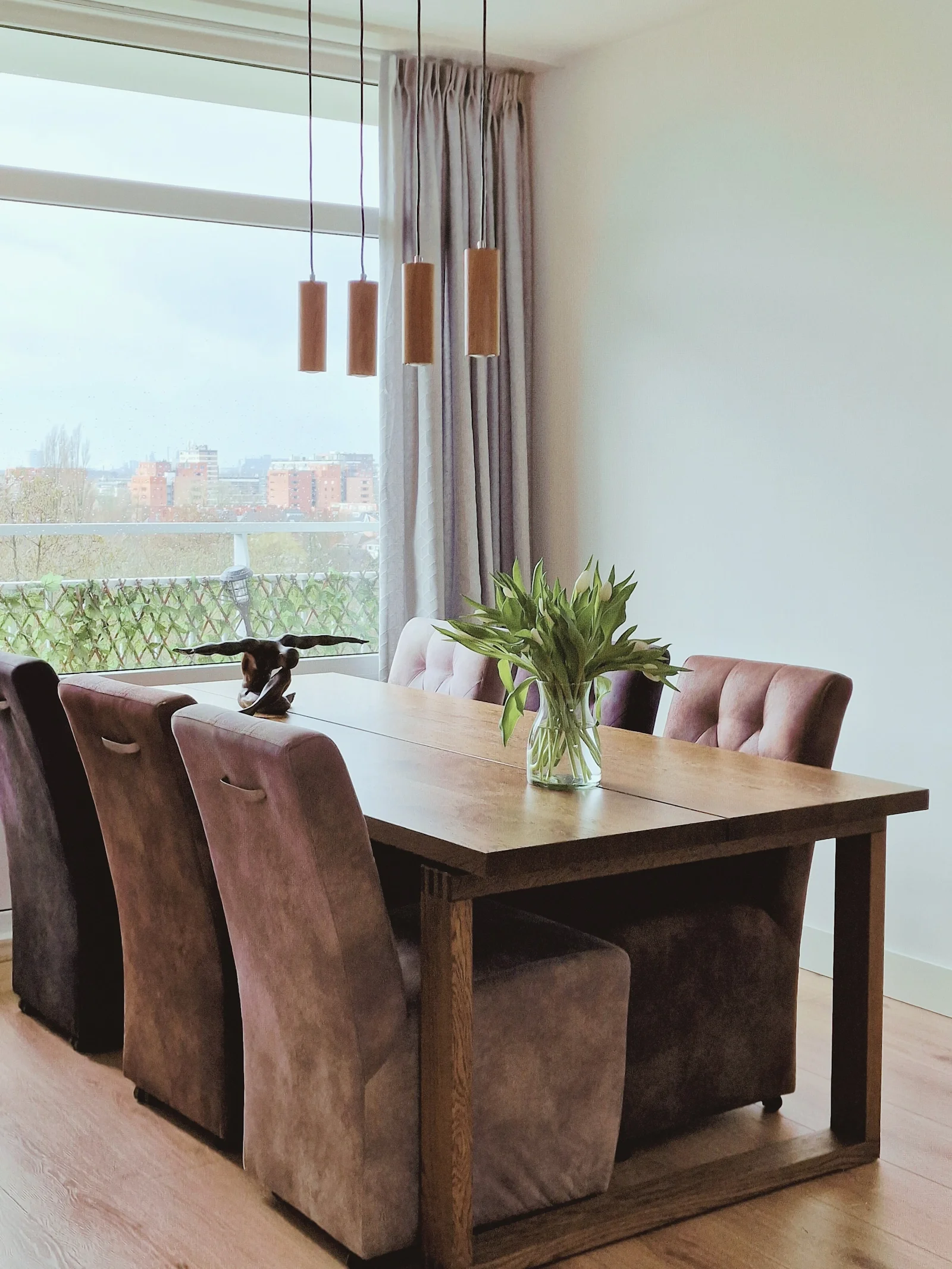 Lichte eetkamer met een houten eettafel, taupe eetkamerstoelen, houten hanglampen en een vaas met tulpen bij het raam met grijze gordijnen.