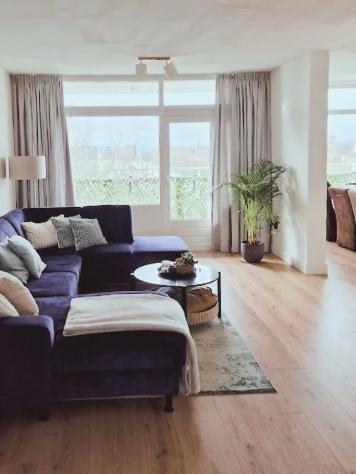 Woonkamer met donkerblauwe hoekbank en ronde salontafel. Neutrale tinten, houtvloer en gordijnen maken het geheel warm en rustig.  Interieuradvies, interieurontwerp en styling door En-Casa Interiors in Noord-Holland.