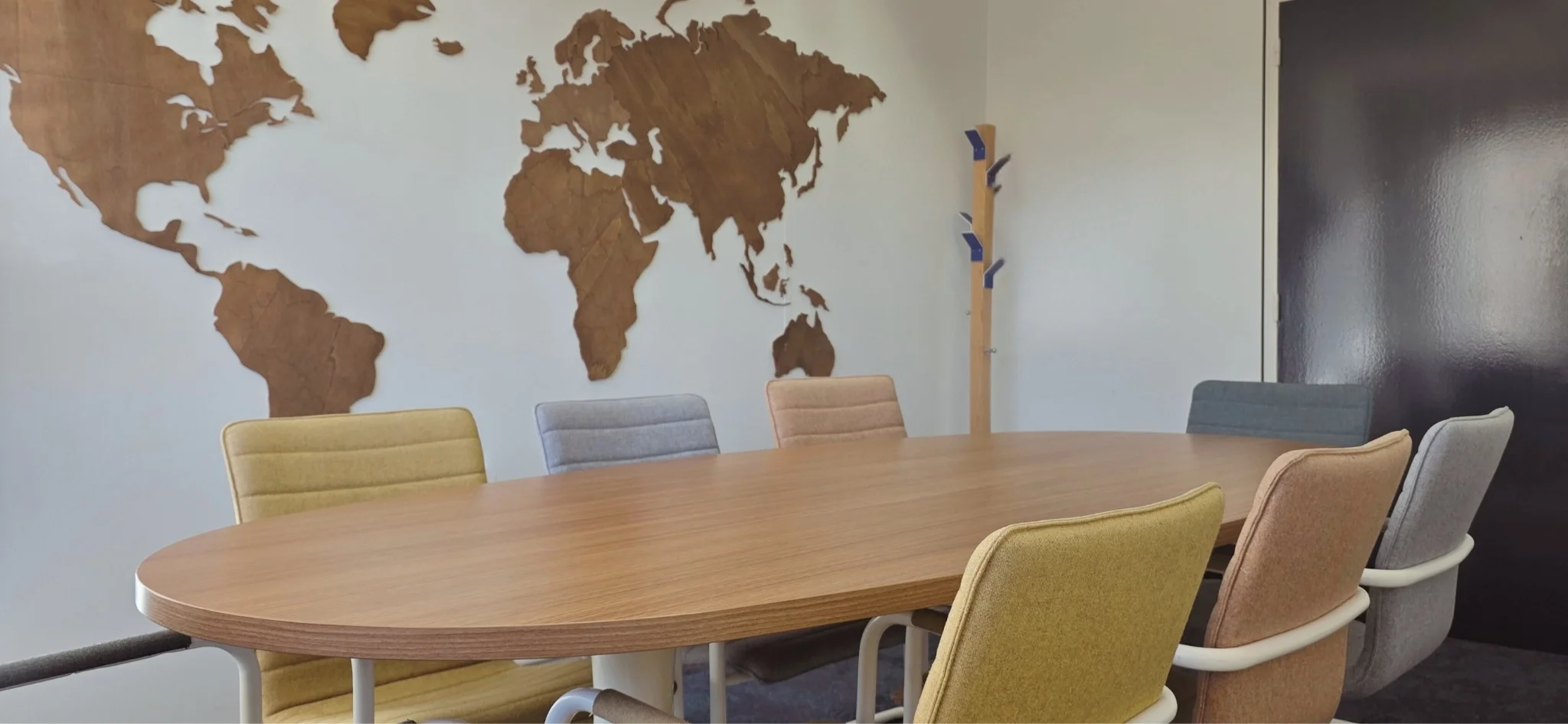 Ovale houten vergadertafel met zachte, kleurrijke stoelen in een rustige vergaderruimte, met een houten wereldkaart als wanddecoratie op de achtergrond.