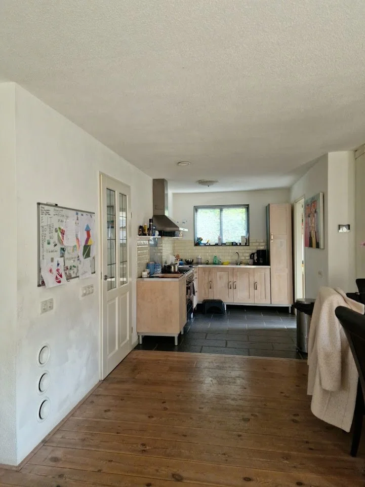 “Keuken in before-situatie met lichte houten keukenkasten, donker tegelvloer en houten vloer in het voorste deel. De keuken heeft een rechte opstelling met afzuigkap, raam boven het aanrecht en zicht op de eetruimte, met een praktische en open indeli