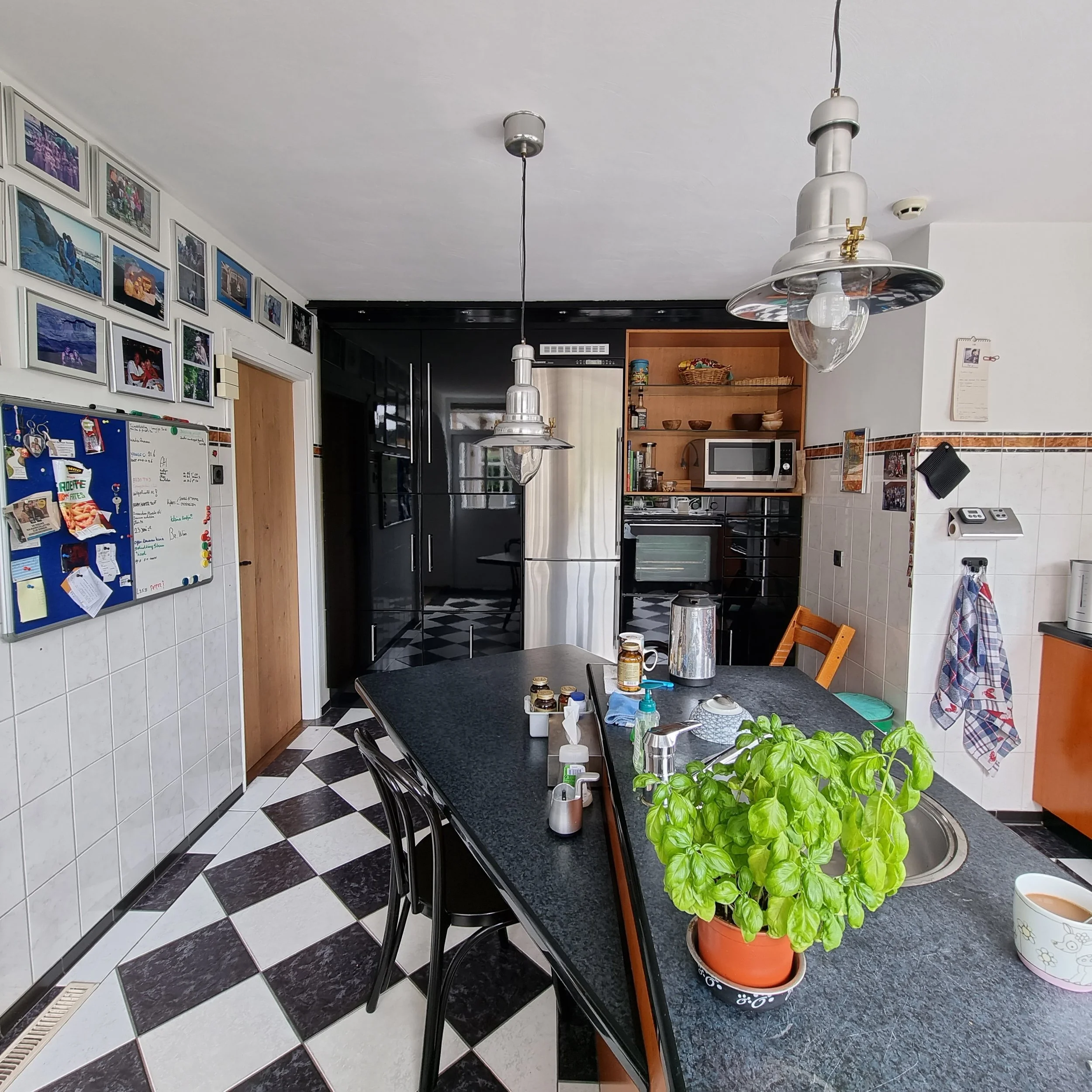 Before foto van de keuken met zwart-witte tegelvloer, donker werkblad en een lang aanrecht met barkrukken. Houten en zwarte kasten, hanglampen en persoonlijke wanddecoratie geven karakter, maar de ruimte oogt nog druk en mist een rustige, moderne een