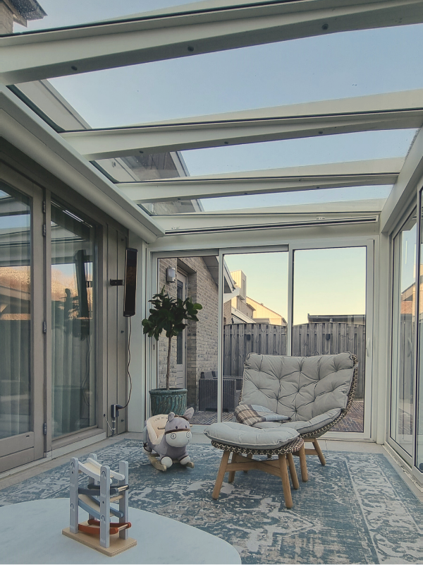Overzicht van de veranda met glazen dak en glazen wanden, ingericht met relaxstoel, vloerkleed en salontafel.
