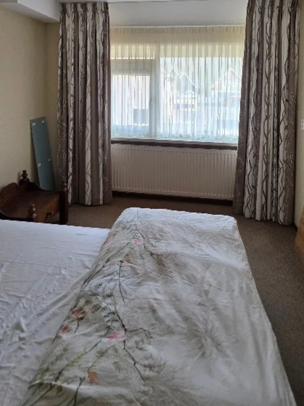 Slaapkamer vóór make-over met raam, gedateerde gordijnen en donkere vloerbedekking – En-Casa nteriors.