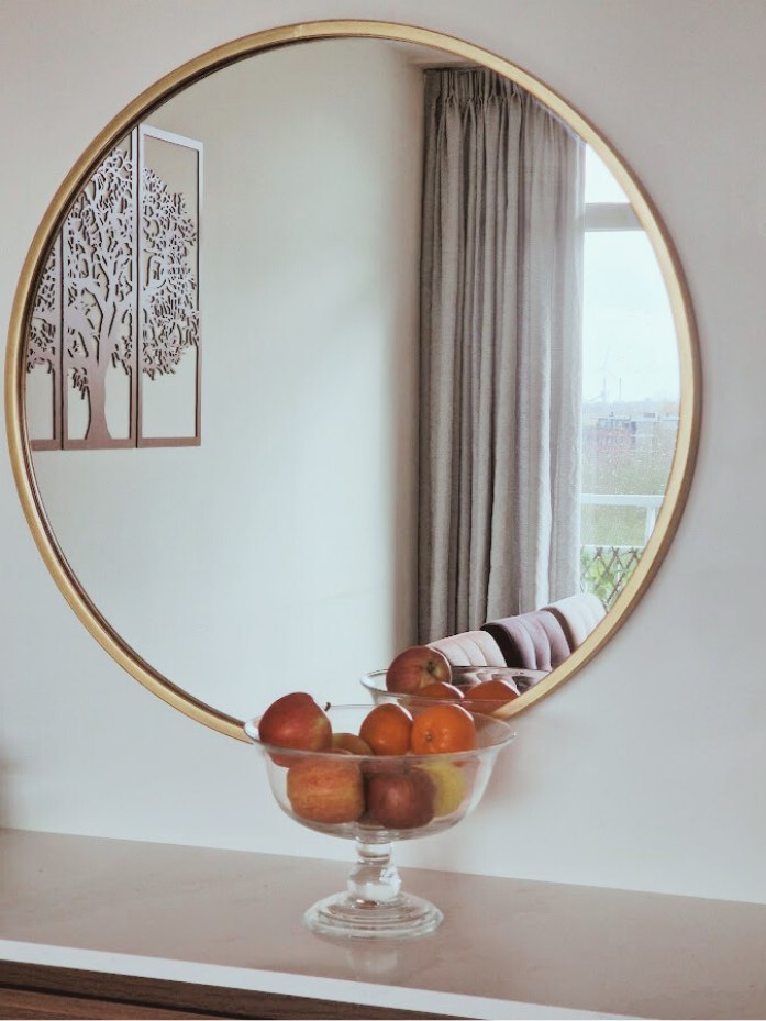 Interieurdetail: ronde spiegel als eyecatcher met subtiele goudtint en een fruitschaal op het dressoir. Minimalistisch, warm en in balans.