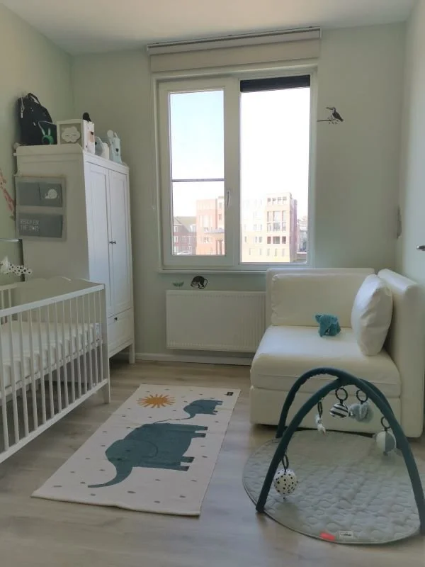 Lichte baby-/kinderkamer met zachtgroene wanden en een houtlook vloer. Links staat een wit ledikant met daarnaast een hoge witte kast. In het midden zorgt het raam met rolgordijn voor veel daglicht, met een radiator eronder. Op de vloer ligt een spee