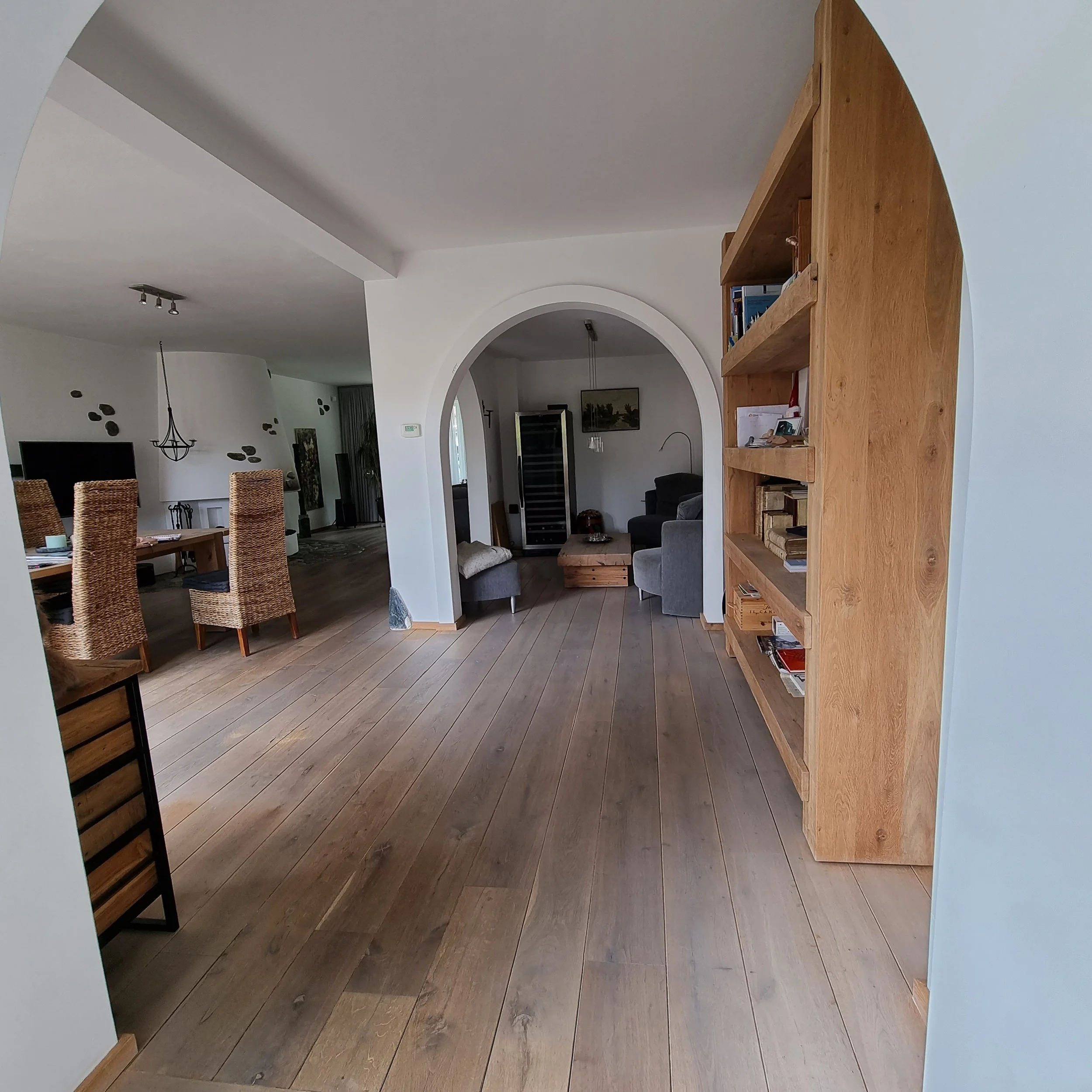 Before foto van de begane grond met lange zichtlijn door de woonruimte, houten vloer en open doorgangen tussen keuken en woonkamer. Maatwerkkast aan de wand en een basisindeling zorgen voor een ruimtelijk gevoel, maar missen nog samenhang en sfeer.