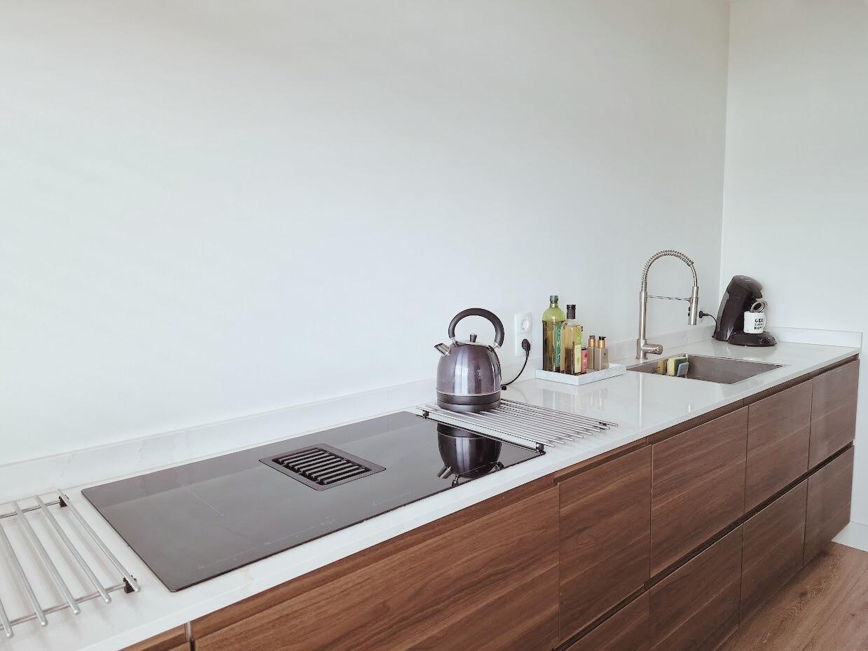 Keuken close-up met lange werkruimte: inductiekookplaat, spoelbak en wit werkblad op warme houtfronten. Strak, rustig en praktisch.