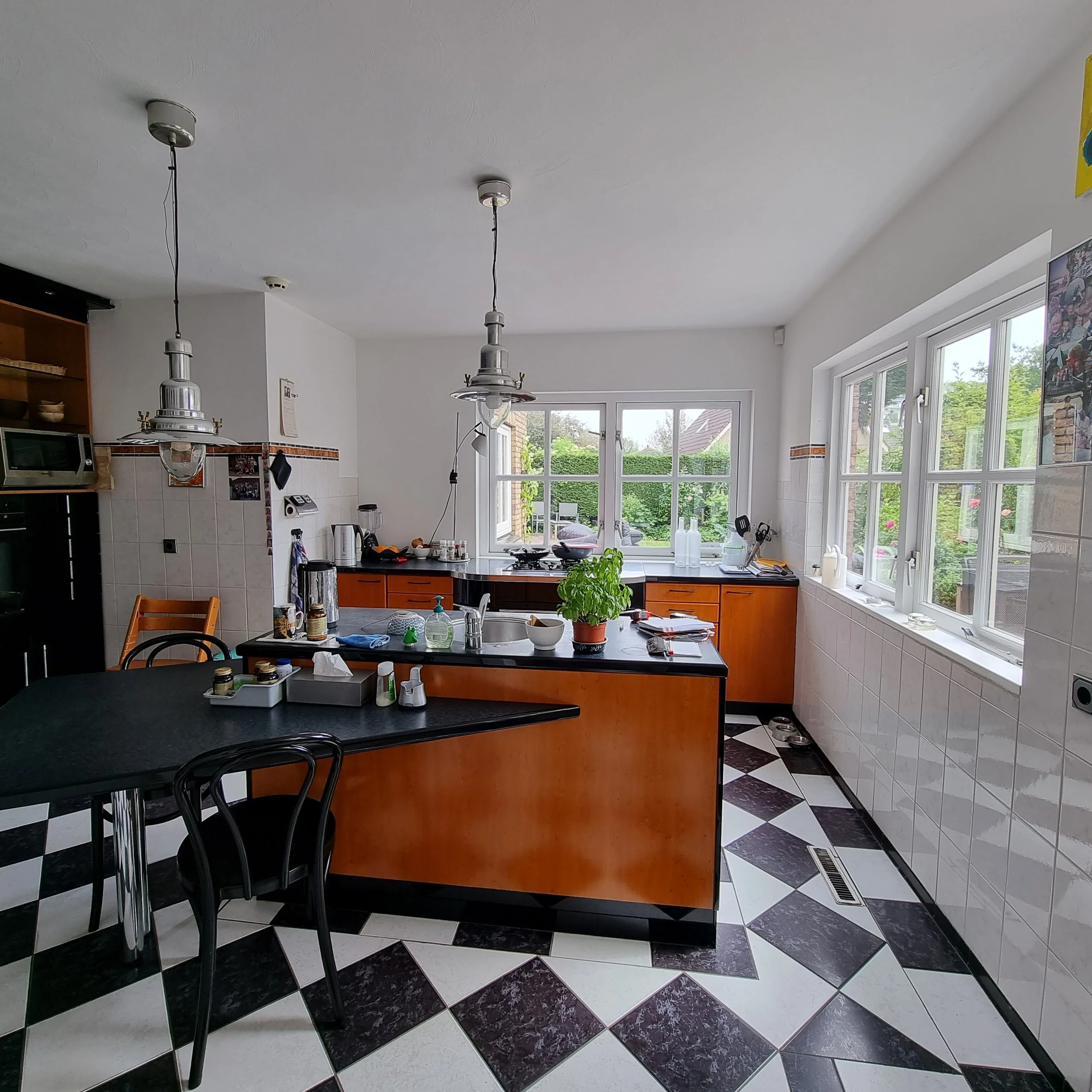 Before foto van de keuken met zwart-witte tegelvloer, houten keukenkasten en een compact kookeiland met barstoelen. Grote ramen zorgen voor veel daglicht en zicht op de tuin, maar de ruimte mist nog een moderne samenhang en rustige basis.