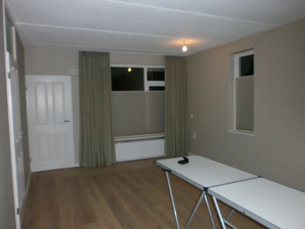 Deze foto toont de voor-situatie van de woonkamer: een lege, rustige ruimte met neutrale wanden, houten vloer en raampartijen met gordijnen. De basis staat al, maar meubels en accessoires ontbreken nog, waardoor de ruimte klaar is om warmte, sfeer en