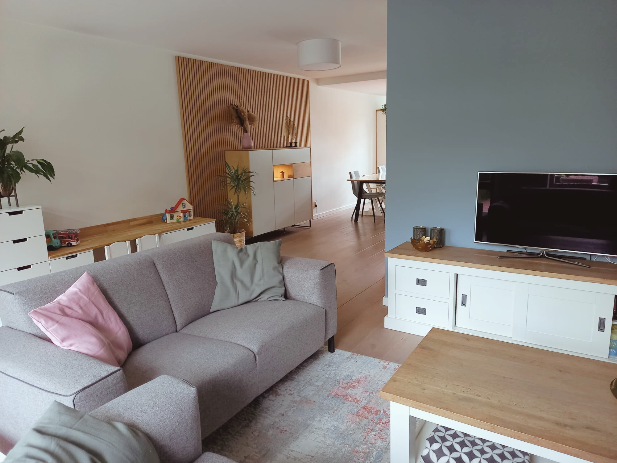 Lichte woonkamer met grijze bank, zachtroze accenten en natuurlijke houttinten. Een rustige, moderne basis met warme details en een open verbinding naar de eethoek — voor een en casa dat fijn voelt en klopt in balans. Ontdek wat En-Casa voor jou kan 