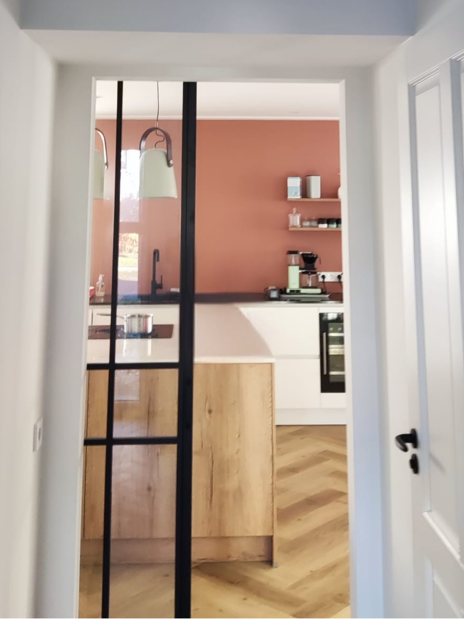Doorkijk vanuit de hal naar de keuken met een stalen glasdeur, een houten kookeiland en een visgraat houten vloer. De keuken combineert witte kasten met een warme terracotta accentwand en natuurlijke materialen, wat zorgt voor een rustige en uitnodig