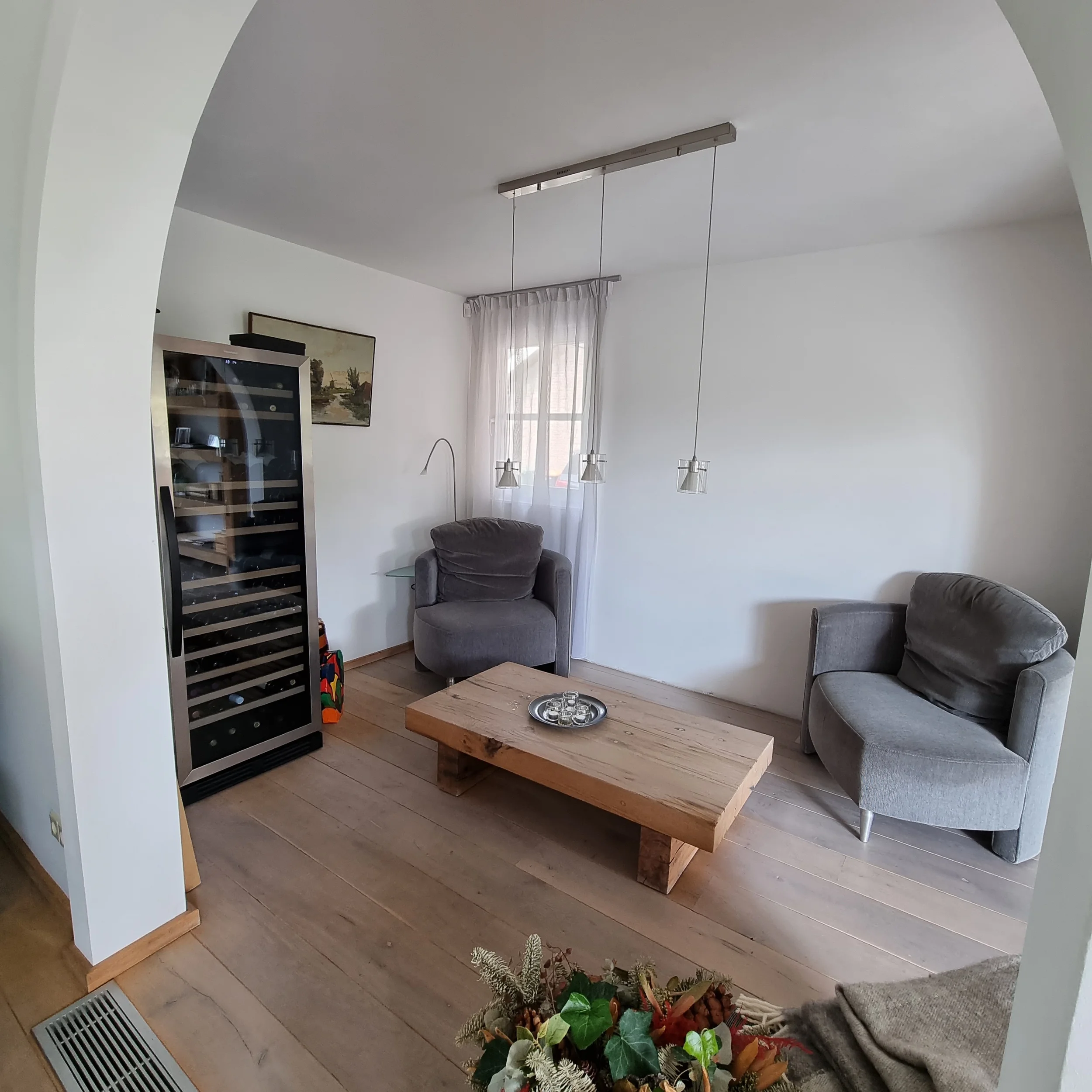 Before foto van een zithoek met houten salontafel, losse fauteuil en witte wanden, gezien door een ronde boogdoorgang. Basisverlichting, een wijnkast en beperkte styling zorgen voor een functionele ruimte met nog weinig warmte en samenhang.