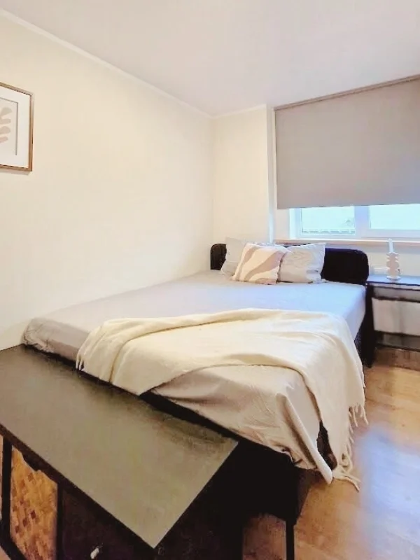 Een rustige slaapkamer met een netjes opgemaakt bed in zachte beige tinten, warme houten vloer en verduisterend rolgordijn — een praktische, serene basis die meteen en casa voelt na een lange dag.