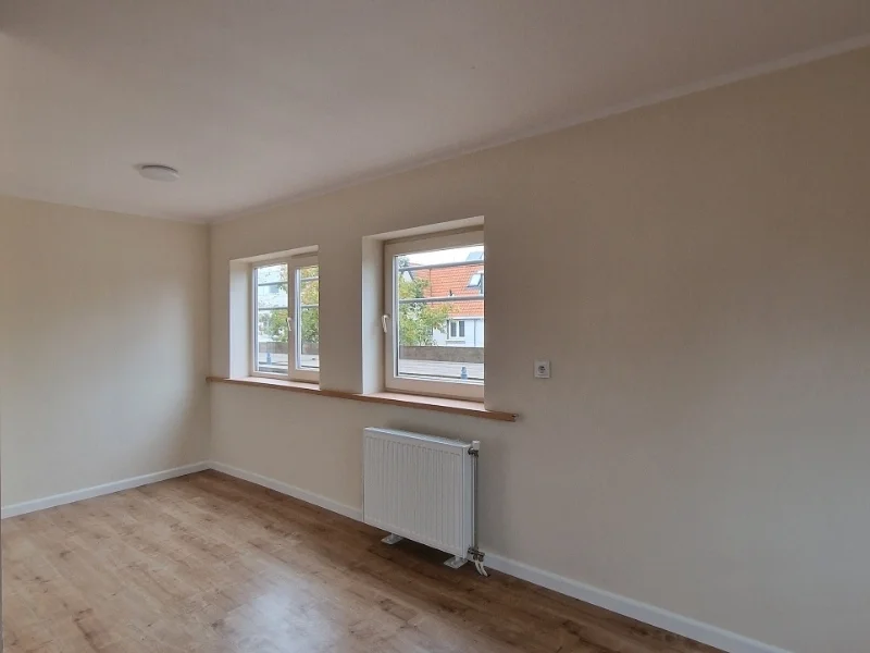 Een lichte “before” kamer met hoekraam, warme houtlook vloer en neutrale wanden — een heldere basis met veel daglicht, perfect om straks een rustige en casa sfeer op te bouwen.