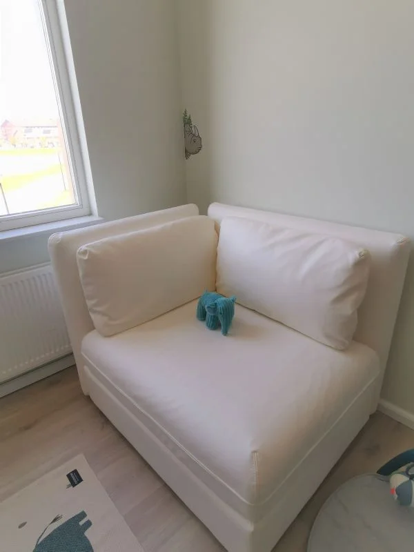Detail van een rustige baby-/kinderkamer: een compact wit bankje/stoel met grote witte kussens staat in de hoek naast het raam. Op de zitting ligt een klein blauw olifantje als decoratie. De muren zijn in een zachte groentint en de vloer heeft een li
