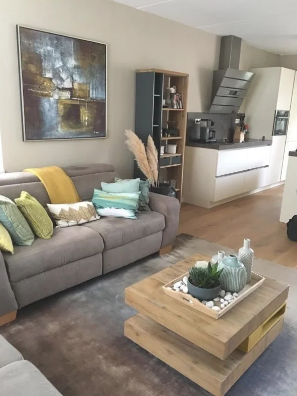 Deze foto toont het totaalbeeld van de woonkamer & eetkamer make-over, met een warme, rustige basis en een fijne open verbinding met de keuken. De taupe bank en het zachte vloerkleed zorgen voor comfort, terwijl het houten salontafelblad warmte en na