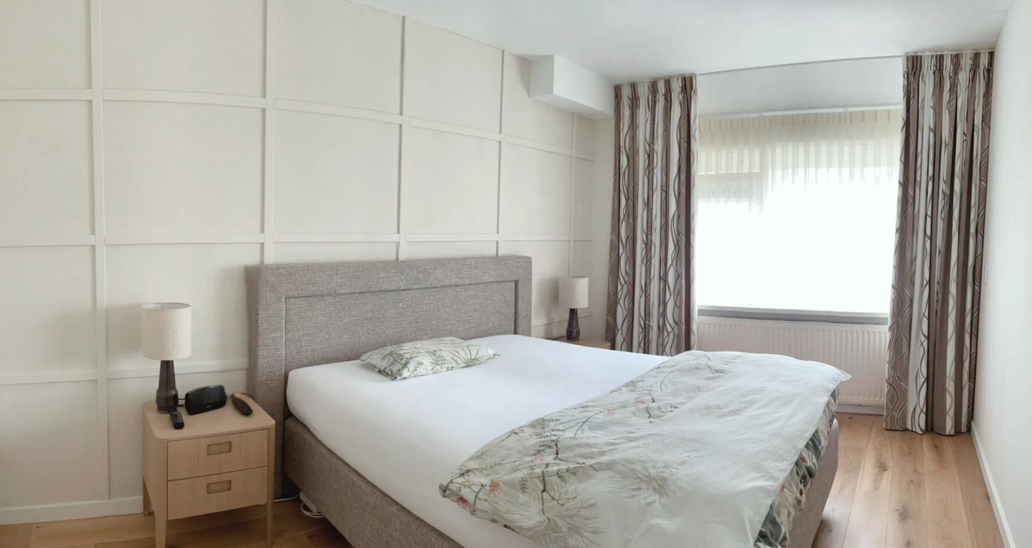 Slaapkamer na make-over met paneelwand, gestoffeerd bed, houten vloer en raam met gordijnen – interieuradvies, interieurontwerp en styling door En-Casa Interiors.