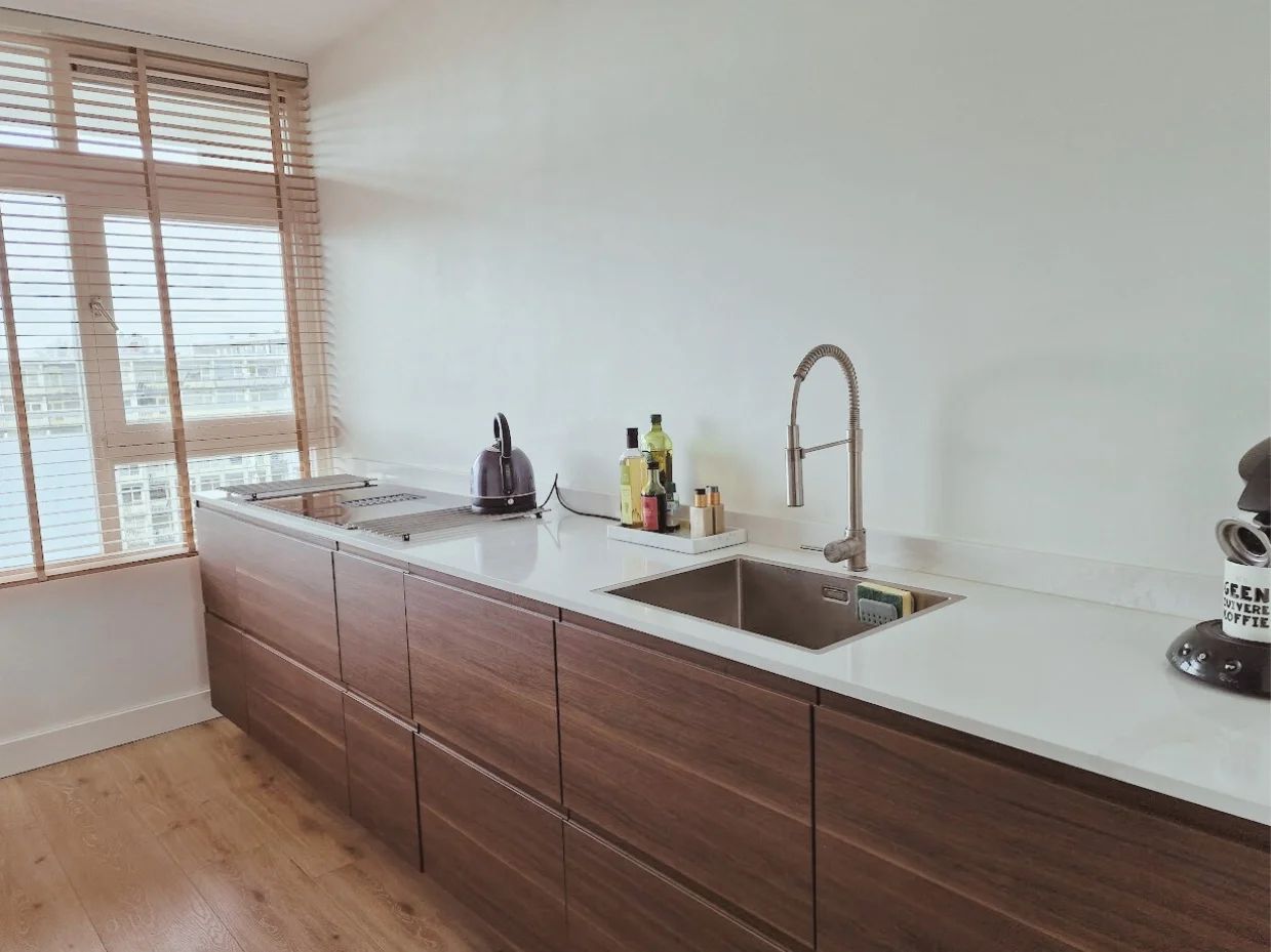 Lange werkruimte in de keuken met warme houtfronten en wit werkblad. Veel licht, strakke lijnen en een rustige, praktische indeling.  Interieuradvies, interieurontwerp en styling door En-Casa Interiors in Noord-Holland.