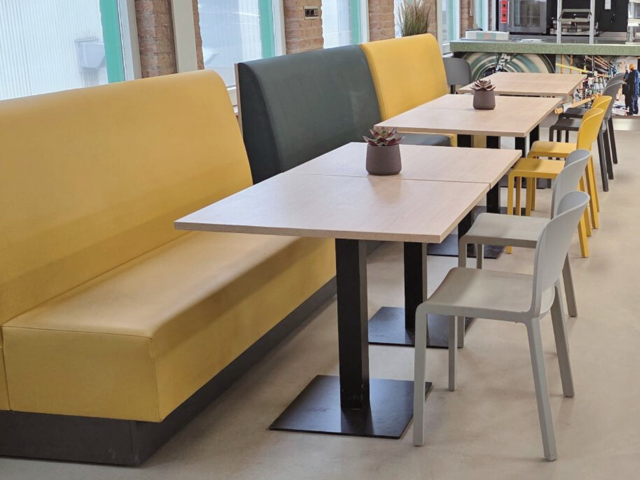 Treinbank-opstelling in warm geel en diep groen, gecombineerd met losse stoelen en strakke tafels, zorgt voor een gezellige en praktische eethoek. De rustige vloer en het slanke zwarte tafelonderstel geven een moderne, industriële basis, terwijl de z