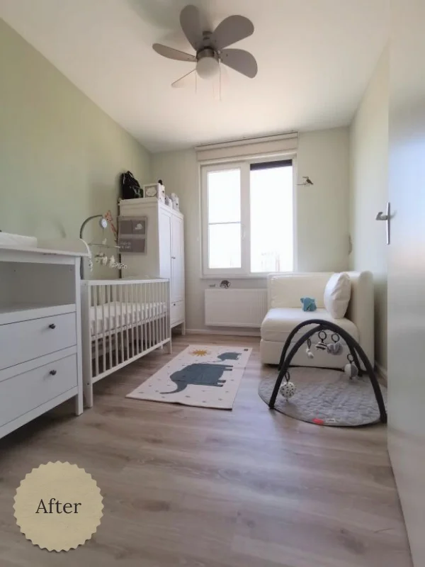 Lichte babykamer met zachtgroene wanden en een warme houtlook vloer. Links staan een witte commode en een wit ledikant. Bij het raam ligt een speels vloerkleed en aan het plafond hangt een ventilator, waardoor de ruimte rustig en luchtig aanvoelt.