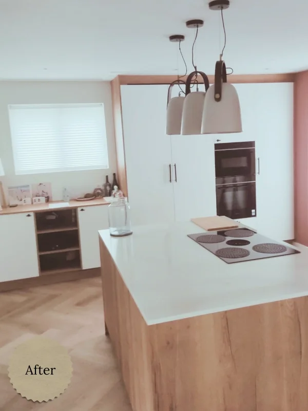 Moderne keuken met een houten kookeiland en wit werkblad, voorzien van een zwarte inductiekookplaat. Boven het eiland hangen drie witte hanglampen met leren details. In de achtergrond zijn witte kasten, een ingebouwde oven en een raam met jaloezieën 