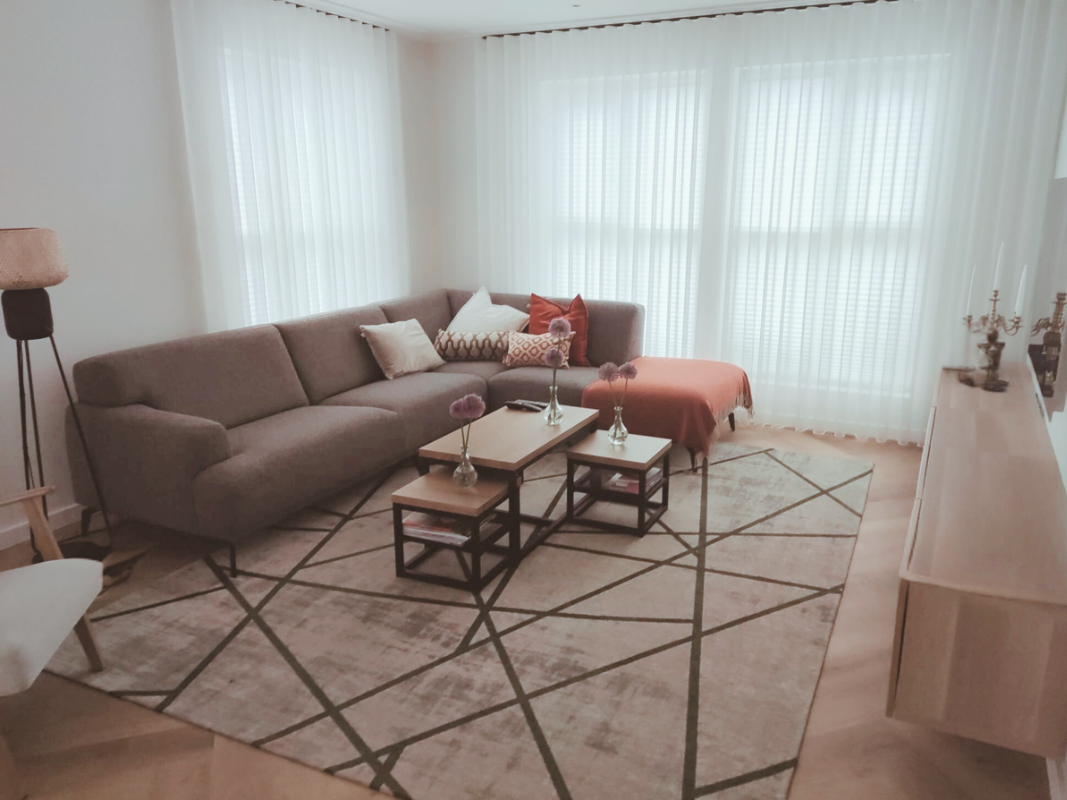 Alt-tekst:
Lichte woonkamer met een ruime hoekbank in zachte beige tinten, aangevuld met kussens in crème en warm terracotta. Voor de bank staan twee houten salontafels met zwarte frames op een groot vloerkleed met grafisch patroon. Grote ramen met t