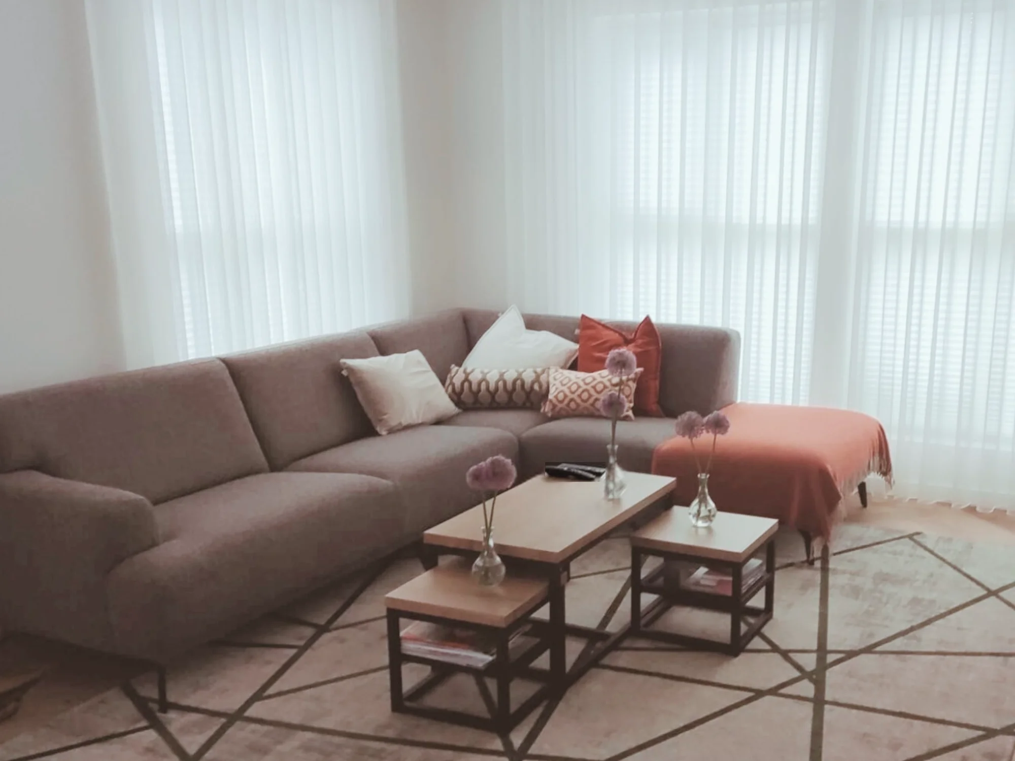 Woonkamer met een ruime hoekbank in zachte beige tinten, aangevuld met kussens in crème en warm terracotta. Voor de bank staan twee houten salontafels met zwarte frames op een groot vloerkleed met grafisch patroon. Grote ramen met transparante gordij